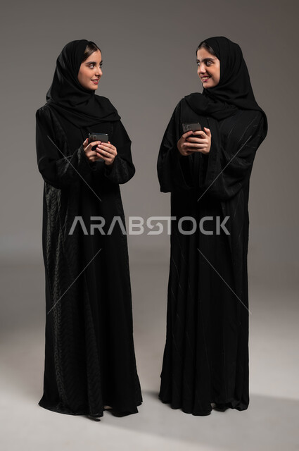 نظرة الأختان لبعضهما البعض بإيماءات السرور، استخدام الأجهزة والتقنيات الحديثة، التواصل الاجتماعي مع الأهل والاصدقاء عبر الموبايل، بورتريه لشابتان عربيتان خليجيتات سعوديتان محجبتان ترتديان عباءة تحملان بيدهما الجوال، صورة الجسم بالطول الكامل، خلفية رمادية