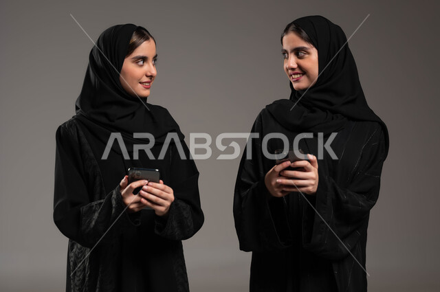 إيماءات الفرح و السعادة، استخدام الأجهزة والتقنيات الحديثة و المتطورة، التواصل الاجتماعي مع الأهل والاصدقاء عبر الموبايل، بورتريه مقرب لشابتان عربيتان خليجيتات سعوديتان محجبتان ترتديان عباءة مبتسمتان تحملان بيدهما الجوال، خلفية رمادية