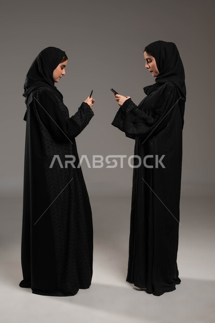 إيماءات الإندماج و التركيز، التواصل الاجتماعي مع الأهل والاصدقاء عبر المحمول، بورتريه لشابتان عربيتان خليجيتات سعوديتان محجبتان ترتديان عباءة تحملان بيديهما الجوال،استخدام الأجهزة والتقنيات الحديثة والمتطورة، صورة لكامل الجسم، خلفية رمادية