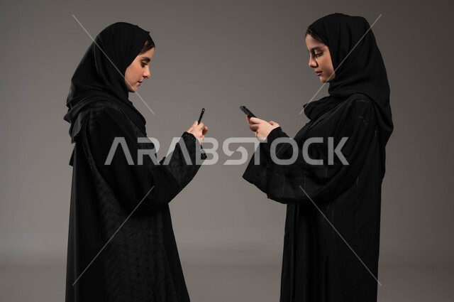 التواصل الاجتماعي مع الاصدقاء عبرالمحمول، بورتريه مقرب لشابتان عربيتان خليجيتات سعوديتان محجبتان ترتديان عباءة تحملان بيديهما الجوال،استخدام الأجهزة والتقنيات الحديثة والمتطورة، الوقوف مقابل بعضهما، ايماءات الاندماج والتركيز، خلفية رمادية