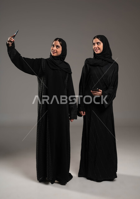 استخدام الأجهزة التقنية المتطورة، توثيق اللحظات السعيدة، بورتريه لشابة عربية خليجية سعودية محجبة ترتدي العباءة السوداء تقوم بالتقاط صور السيلفي التذكارية مع اختها التوءم بإيماءات السعادة والسرور، صورة الجسم بالكامل، خلفية رمادية
