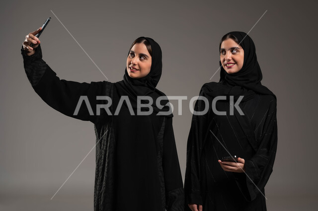 توثيق اللحظات السعيدة، استخدام الأجهزة التقنية المتطورة، بورتريه مقرب لشابة عربية خليجية سعودية محجبة ترتدي العباءة السوداء تقوم بالتقاط صور السيلفي التذكارية مع اختها التوءم بإيماءات السعادة والسرور، خلفية رمادية