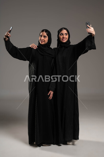 Documenting happy moments, using advanced devices and technologies, a portrait of two veiled Gulf Arab young women wearing the black abaya, taking souvenir selfies with gestures of happiness and pleasure, full body portrait, gray background