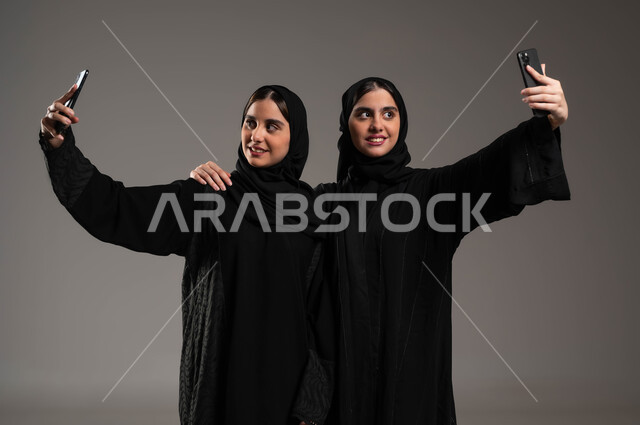 الاستعداد لالتقاط صور تذكارية عبر الجوال، النظر الى كاميره الموبايل بإيماءات السرور و السعادة، بورتريه مقرب لبنتان توءم عربيتان سعوديتان خليجيتان محجبتان ترتديان عباءة سوداء تقومان باخد صورة باستعمال الهاتف المحمول، استخدام التكنولوجيا الحديثة، خلفية رمادية
