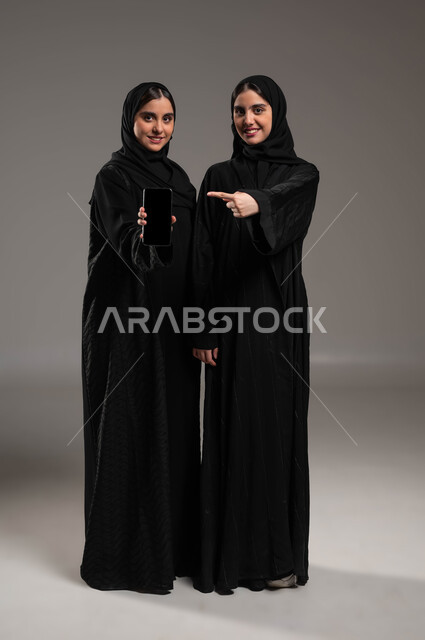 استخدام التكنولوجيا الحديثة، عرض تطبيقات وخدمات الهاتف المحمول، بورتريه لفتاتان عربيتان خليجيتان سعوديتان مبتسمتان ترتديان عباءة سوادء إحداهما تحمل بيدها جوال بشاشه سوداء فارغة و الاخرى تشير بيدها إلى الموبايل، صورة الجسم بالكامل، خلفيه رماديه