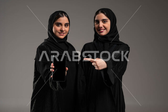 عرض تطبيقات وخدمات الهاتف المحمول، بورتريه مقرب لفتاتان عربيتان خليجيتان سعوديتان مبتسمتان ترتديان عباءة سوادء إحداهما تحمل بيدها جوال بشاشه سوداء فارغة و الاخرى تشير بيدها إلى الهاتف ،النظر إلى الكاميرا بإيماءات السرور، استخدام التكنولوجيا الحديثة، خلفيه رماديه