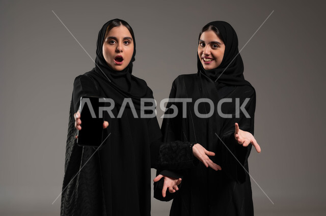 إيماءات الدهشة والتساؤل والاستغراب، عرض تطبيقات وخدمات الهاتف المحمول، بورتريه مقرب لفتاتان عربيتان خليجيتان سعوديتان مبتسمتان ترتديان عباءة سوداء، النظر إلى الكاميرا بتعابير السرور، استخدام التكنولوجيا الحديثة، خلفيه رماديه