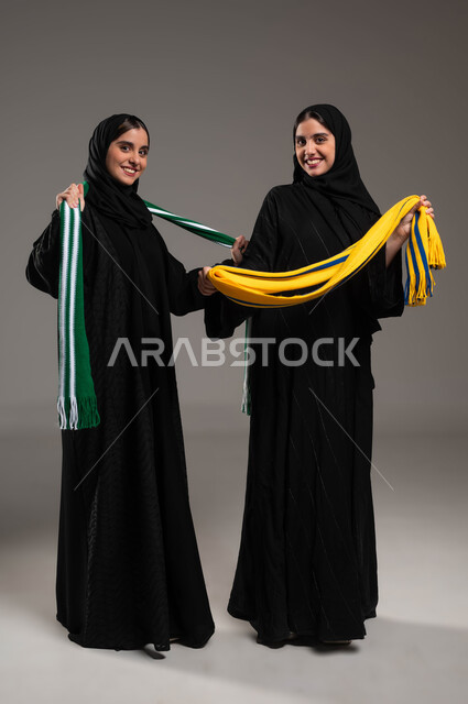 الفوز و الانتصار بالمباراة، الشغف بالأنشطة الترفيهية، المباريات والألعاب الرياضية، تعابير الفرح  والسرور لانتصار الفريق، بورتريه مقرب لبنتان توءم عربيتان خليجيتان سعوديتان ترتديان أوشحة تشجيعية لفرق كرة القدم تقومان بالتباهي بفريقهما، خلفية رمادية