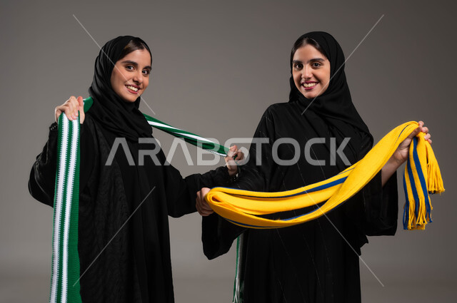 الشغف بالأنشطة الترفيهية، النظر الى الكاميرا بإيماءات الحماس والشغف، المباريات والألعاب الرياضية، تعابير الفرح  والسرور لانتصار الفريق، بورتريه مقرب لبنتان توءم عربيتان خليجيتان سعوديتان ترتديان أوشحة تشجيعية لفرق كرة القدم تقومان بالتباهي بفريقهما، خلفية رمادية