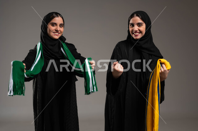 أنشطة رياضية ترفيهية، النظر الى الكاميرا بإيماءات الحماس و الشغف، المباريات والألعاب الرياضية، تعابير الفرح  والسرور لانتصار الفريق، بورتريه مقرب لبنتان توءم عربيتان خليجيتان سعوديتان ترتديان أوشحة تشجيعية لفرق كرة القدم تقومان بالتباهي بفريقهما، خلفية رمادية