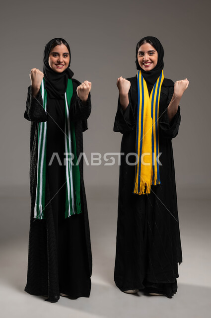 رفع اليدين بإيماءات الفوز ، النظر الى الكاميرا بإيماءات الحماس و الشغف، المباريات والألعاب الرياضية، تعابير الفرح  والسرور لانتصار الفريق، بورتريه لبنتان توءم عربيتان خليجيتان سعوديتان ترتديان أوشحة تشجيعية لفرق كرة القدم، صورة لكامل الجسم، خلفية رمادية