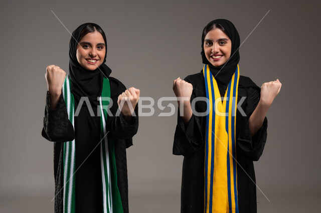 Looking at the camera with gestures of enthusiasm and passion, matches and sports, expressions of joy and pleasure for the victory of the team, a portrait close to two Saudi twins, two Saudi Gulf Arabs wearing scarves of encouragement for football teams, raising hands with winning expressions, gray background