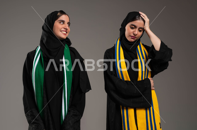 تعابير الفرح بالنصر والفوز، إيماءات الحزن لخسارة الفريق، بورتريه مقرب لفتاة عربية خليجية سعودية محجبة تقوم بإغاظة أختها التوأم لانتصار فريقها المفضل، ارتداء اوشحة تشجيعية لفرق كرة القدم، المباريات والألعاب الرياضية، خلفية رمادية
