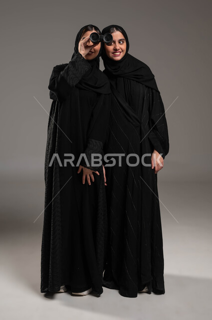 مفهوم المشاركة والاستكشاف والمغامرة، طموحات وأهداف مستقبلية، البحث باستخدام الناظور، بورتريه مقرب لتوأم بنات عربيات خليجيات سعوديات محجبات تتشاركان في النظرعبر المنظار ثنائي العينين، صورة الجسم بالطول الكامل، خلفية رمادية