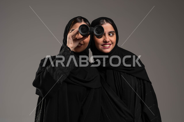 مفهوم الاستكشاف والمغامرة، طموحات وأهداف مستقبلية، البحث باستخدام الناظور، بورتريه مقرب لتوأم بنات عربيات خليجيات سعوديات محجبات تتشاركان في النظر عبر المنظار ثنائي العينين، خلفية رمادية
