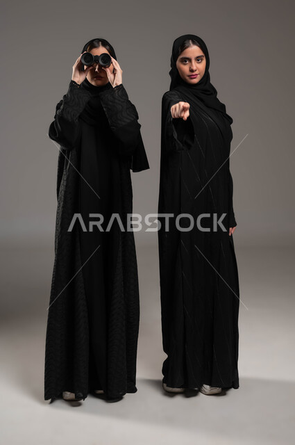 Research using binoculars, the concept of exploration and adventure, future goals and ambitions, a portrait of twin veiled Saudi Gulf Arab girls, one looking through binoculars and the other pointing with her finger to the camera, full-length body portrait, gray backgroun