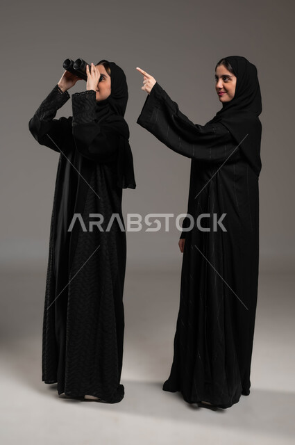 أهداف وطموحات مستقبلية، البحث باستخدام الناظور، بورتريه لتوأم بنات عربيات خليجيات سعوديات محجبات إحداهما تنظر عبر المنظار ثنائي العينين والأخرى تشير بإصبعها إلى شيء ما، مفهوم الاستكشاف والمغامرة، صورة الجسم بالطول الكامل، خلفية رمادية