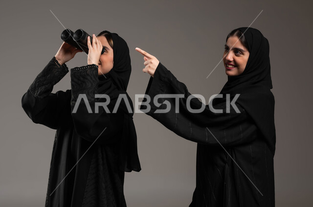 طموحات وأهداف مستقبلية، البحث باستخدام الناظور، بورتريه لتوأم بنات عربيات خليجيات سعوديات محجبات إحداهما تنظر عبر المنظار ثنائي العينين والأخرى تشير بإصبعها إلى شيء ما، مفهوم الاستكشاف والمغامرة، خلفية رمادية
