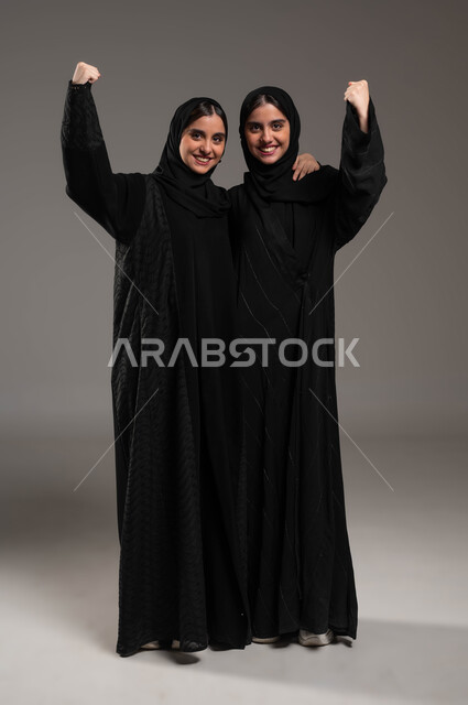 الشعور بالفرح والسرور، مشاعر المحبة والود بين الاخوة، بورتريه لفتاة عربية خليجية سعودية مبتسمة محجبة ترتدي العباءة السوداء تعانق اختها التوأم، رفع اليدين عاليا بإيماءات الفوز والنجاح وتحقيق الأهداف، صورة الجسم بالطول الكامل، خلفية رمادية