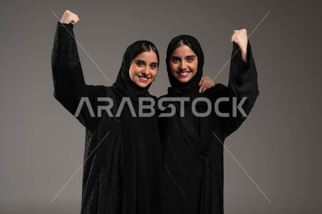 Feelings of love and friendliness between brothers, a feeling of joy and happiness, a close-up portrait of a smiling, veiled Saudi Arabian Gulf girl wearing a black abaya hugging her twin sister, raising hands high with gestures of victory, success and achieving goals, gray backgroun