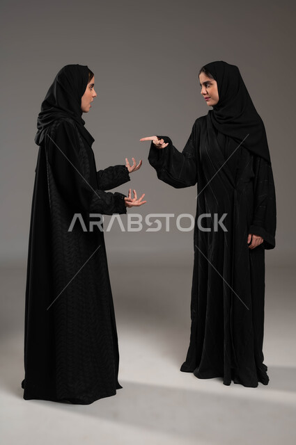 تعابير التعصب والتوتر الانفعالي، مفهوم النقاش الحاد والمواجهة بين الأخوات، بورتريه لتوأم بنات عربيات خليجيات سعوديات محجبات تقفان مقابل بعضهما وتتجادلان بإيماءات الغضب وإلقاء اللوم، صورة الجسم بالطول الكامل، خلفية رمادية