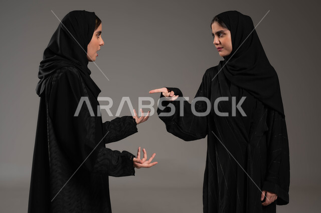 مفهوم النقاش الحاد والمواجهة بين الأخوات، بورتريه مقرب لتوأم بنات عربيات خليجيات سعوديات محجبات تقفان مقابل بعضهما وتتجادلان بإيماءات الغضب وإلقاء اللوم، تعابير التعصب والتوتر الانفعالي، خلفية رمادية