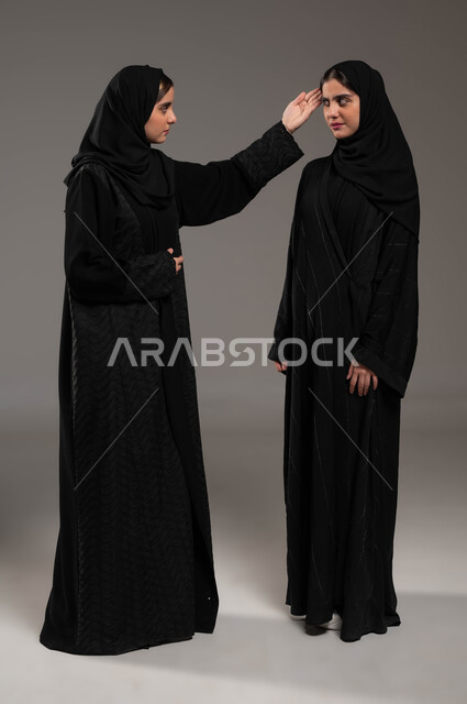 Asking for focus and logical thinking, reprimanding, blaming and admonishing, a portrait of a veiled Saudi Gulf Arab woman wearing a black abaya, placing her palm on her twin sister’s head, gestures of anger, fanaticism and emotional tension, full-length body image, gray background