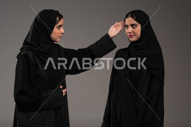 التوبيخ واللوم والمعاتبه، بورتريه مقرب لشابه عربيه خليجية سعودية محجبة ترتدي العباءة السوداء تضع كفها على راس اختها التوام ، طلب التركيز والتفكير المنطقي، إيماءات الغضب والتعصب والتوتر الانفعالي، مفهوم الجدال الحاد والمواجهة بين الأخوات، خلفية رمادية