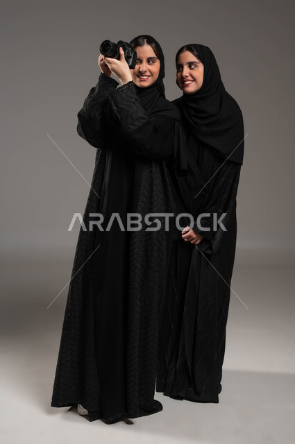 الاحتراف والموهبة في استخدام آلة التصوير الفوتوغرافي، توثيق اللحظات السعيدة، بورتريه لتوأم بنات عربيات خليجيات سعوديات محجبات ترتديان العباءة السوداء تستمتعان بالتقاط الصور في الكاميرا، صورة الجسم بالطول الكامل، خلفية رمادية