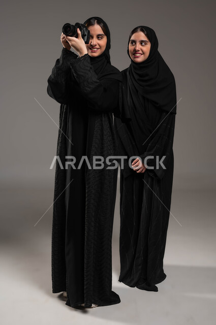 الموهبة والاحتراف في استخدام آلة التصوير الفوتوغرافي، توثيق اللحظات السعيدة، بورتريه لتوأم بنات عربيات خليجيات سعوديات محجبات ترتديان العباءة السوداء تستمتعان بالتقاط الصور في الكاميرا، صورة الجسم بالطول الكامل، خلفية رمادية