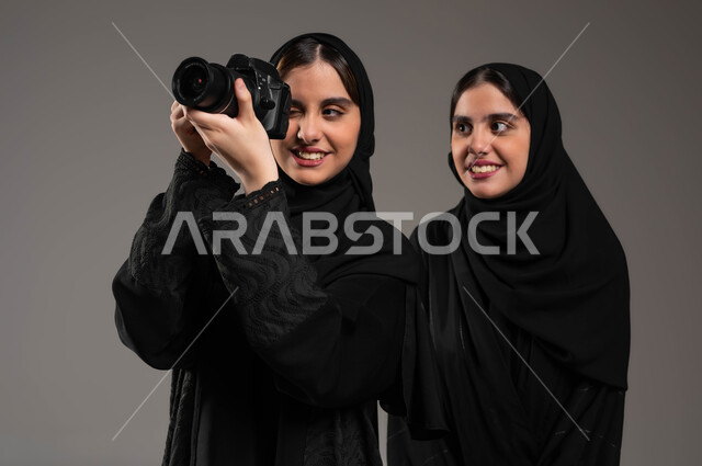 The concept of talent and professionalism, using a camera to document happy moments, a close-up portrait of twin veiled Saudi Gulf Arab girls wearing the black abaya enjoying taking pictures with the camera, gray backgroun