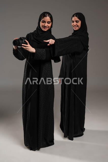 الإشارة إلى اللقطات المتميزة، فنون وهوايات التصوير الفوتوغرافي، بورتريه لتوأم بنات عربيات خليجيات سعوديات محجبات ترتديان العباءة السوداء تستمتعان بالنظر الى الصور في الكاميرا، توثيق اللحظات السعيدة، صورة الجسم الطول الكامل، خلفية رمادية