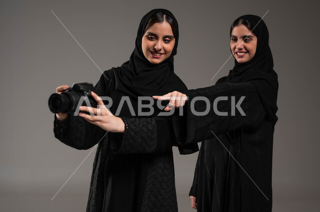 الإشاره إلى اللقطات المتميزة بإيماءات السرور، فنون وهوايات التصوير الفوتوغرافي، بورتريه لتوأم بنات عربيات خليجيات سعوديات محجبات ترتديان العباءة السوداء تستمتعان بالنظر الى الصور في الكاميرا، توثيق اللحظات السعيدة، خلفية رمادية