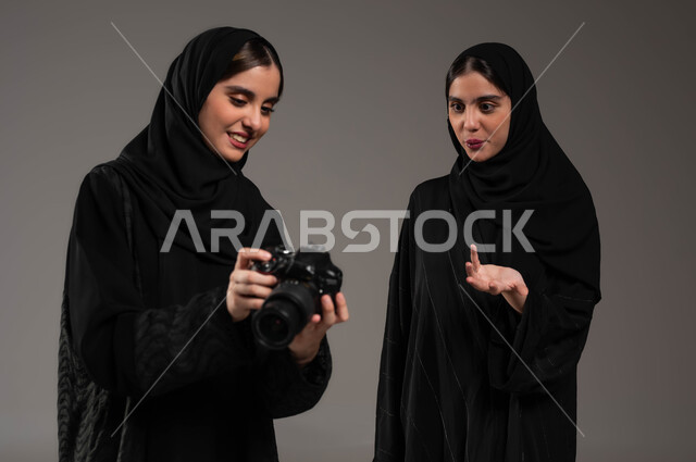 هوايات و فنون التصوير الفوتوغرافي، بورتريه لتوأم بنات عربيات خليجيات سعوديات محجبات ترتديان العباءة السوداء تستمتعان بالنظر الى الصور في الكاميرا، إيماءات التعجب والذهول، توثيق اللحظات السعيدة، خلفية رمادية