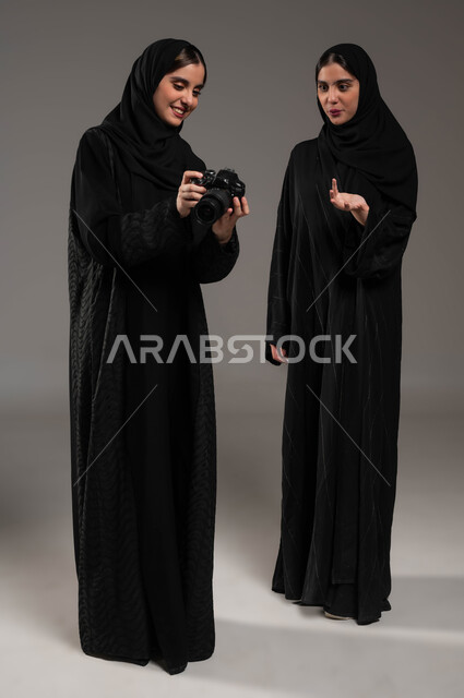 إيماءات التعجب والذهول، هوايات و فنون التصوير الفوتوغرافي، بورتريه لتوأم بنات عربيات خليجيات سعوديات محجبات ترتديان العباءة السوداء تستمتعان بالنظر الى الصور في الكاميرا، توثيق اللحظات السعيدة، صورة الجسم الطول الكامل، خلفية رمادية