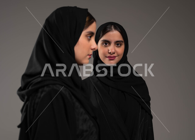 التحديق إلى شيء ما بإيماءات التركيز، بورتريه مقرب لتوأم بنات عربيات خليجيات سعوديات محجبات إحداهما تنظر الى الجانب  والاخرى تنظر إلى الكاميرا بابتسامة، ارتداء العباءة الخليجية السوداء، مفهوم التأمل والتخطيط، خلفية رمادية