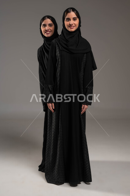 النظر إلى الكاميرا بإيماءات الفرح، بورتريه لفتاة عربية خليجية سعودية محجبة مبتسمة ترتدي العباءة السوداء تقف خلف أختها التوأم، الشعور بالسعادة والسرور، التشارك واللعب مع الأخوات، قضاء أوقات ممتعة ومسلية، صورة الجسم بالطول الكامل، خلفية رمادية