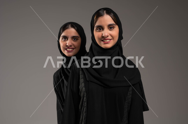 الشعور بالسعادة والسرور، بورتريه مقرب لفتاة عربية خليجية سعودية محجبة مبتسمة ترتدي العباءة السوداء تقف خلف أختها التوأم، النظر إلى الكاميرا بإيماءات الفرح، التشارك واللعب مع الأخوات، قضاء أوقات ممتعة ومسلية، خلفية رمادية