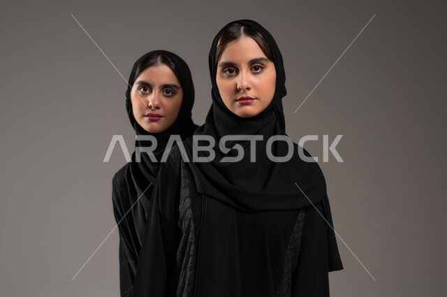 التحديق إلى شيء ما بإيماءات الإحباط، بورتريه لفتاة عربية خليجية سعودية محجبة ترتدي العباءة السوداء تقف خلف أختها تنظران إلى الكاميرا بإيماءات الحزن، الشعور بالخسارة والفشل، خلفية رمادية