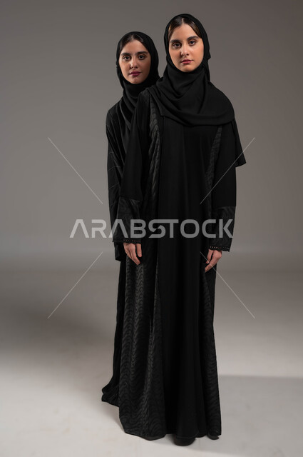 التحديق إلى شيء ما بإيماءات الإحباط، بورتريه لفتاة عربية خليجية سعودية محجبة ترتدي العباءة السوداء تقف خلف أختها تنظران إلى الكاميرا بإيماءات الحزن، الشعور بالخسارة والفشل، صورة الجسم بالطول الكامل, خلفية رمادية