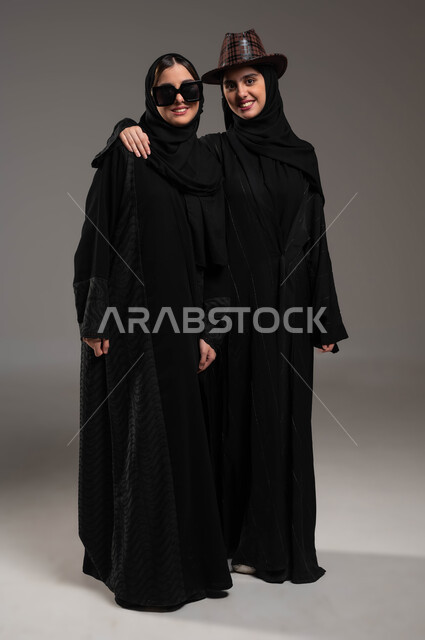 الاستمتاع بقضاء أوقات ممتعة, المحبة والود بين الاخوة، بورتريه لبنتان توءم عربيتان خليجيتان سعوديتان ترتديان العباءة والحجاب تنظران إلى الكاميرا, ارتداء النظارة الشمسية والقبعة التتكرية، صورة الجسم بالطول الكامل، خلفية رمادية