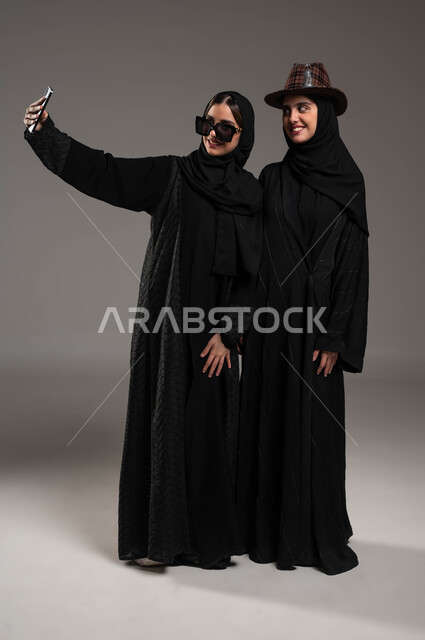 التقاط صور السيلفي التذكارية بايماءات الفرح والسرور, بورتريه لبنتان توءم عربيتان خليجيتان سعوديتان أحدهما تضع قبعة, استخدام الأجهزة والتقنيات الحديثة, توثيق اللحظات السعيدة عبر الهاتف المحمول, صورة الجسم بالطول الكامل، خلفية رمادية