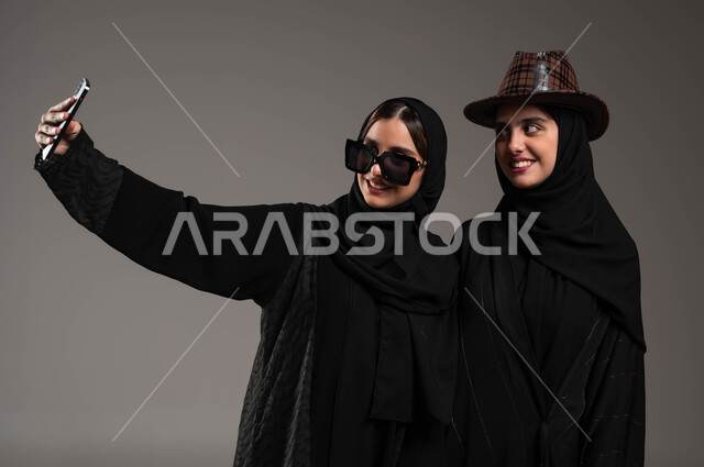 توثيق اللحظات السعيدة عبر الهاتف المحمول, بورتريه مقرب لبنتان توءم عربيتان خليجيتان سعوديتان أحدهما تضع قبعة, استخدام الأجهزة والتقنيات الحديثة, التقاط صور السيلفي التذكارية بايماءات الفرح والسرور, خلفية رمادية