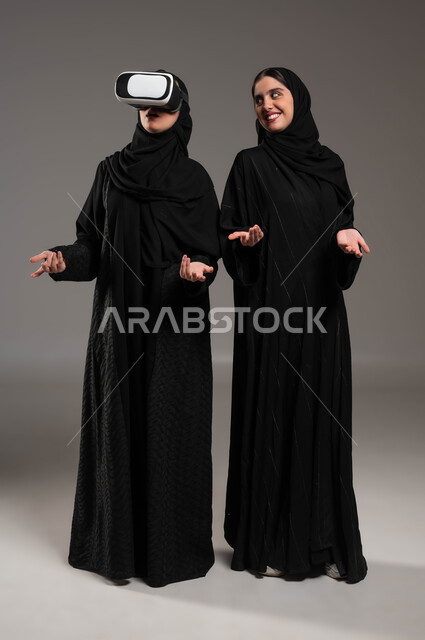 قضاء أوقات ممتعة ومسلية بألعاب الواقع الافتراضي, بورتريه لتوأم بنات عربيات خليجيات سعوديات ترتديان العباءة والحجاب أحدهما ترتدي نظارة الواقع الافتراضي ثلاثية الأبعاد, استخدام الأجهزة التقنية المتطورة بايماءات الفرح والحماس والسرور, صورة الجسم بالطول الكامل، خلفية رمادية