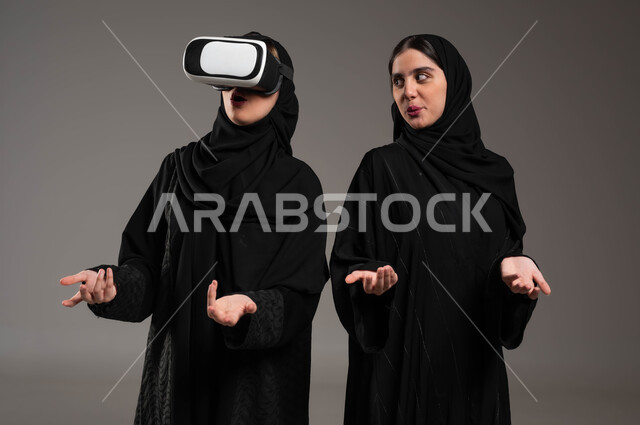 Advanced technologies and devices, simulating the imaginary world, spending fun and entertaining times with virtual reality games, close-up portrait of twin girls, two Saudi Gulf Arab women wearing the abaya and hijab, one of whom is wearing 3D virtual reality glasses, using advanced technical devices with gestures of amazement and astonishment, gray background