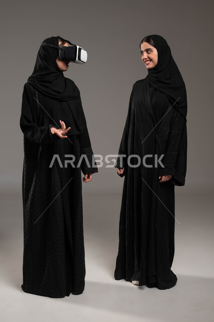 قضاء أوقات ممتعة ومسلية بالألعاب الافتراضية، بورتريه لبنتان توءم عربيتان سعوديتان خليجيتان ترتديان الحجاب والعباءة احداهما ترتدي نظارة ثلاثية الأبعاد,الاستمتاع بمشاهدة أفلام الواقع الافتراضي، محاكاة العالم الخيالي، صورة الجسم بالطول الكامل، خلفية رمادية