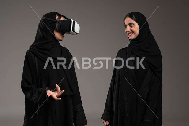 الاستمتاع بمشاهدة أفلام الواقع الافتراضي، قضاء أوقات ممتعة ومسلية بالألعاب الافتراضية، بورتريه مقرب لبنتان توءم عربيتان سعوديتان خليجيتان ترتديان الحجاب والعباءة احداهما ترتدي نظارة ثلاثية الأبعاد، محاكاة العالم الخيالي,  خلفية رمادية