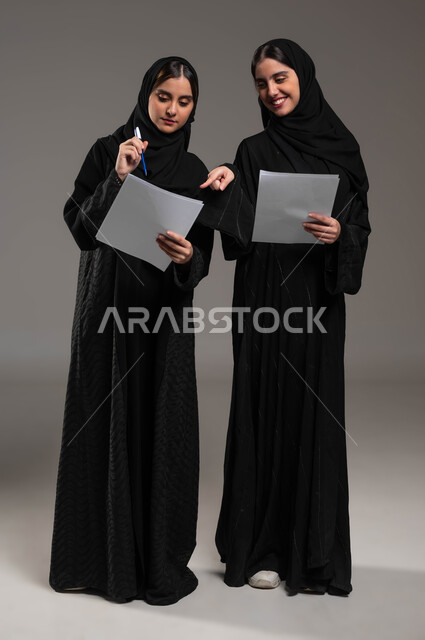 مناقشة الأعمال مع زملاء العمل، المشاركة والتعاون في إنجاز العمل، ايماءات الفرح والسعادة,  الاشارة بالاصبع على أوراق العمل, بورتريه لبنتان عربيتان سعوديتان خليجيتان ترتديان الحجاب والعباءة، صورة الجسم بالطول الكامل، خلفية رمادية