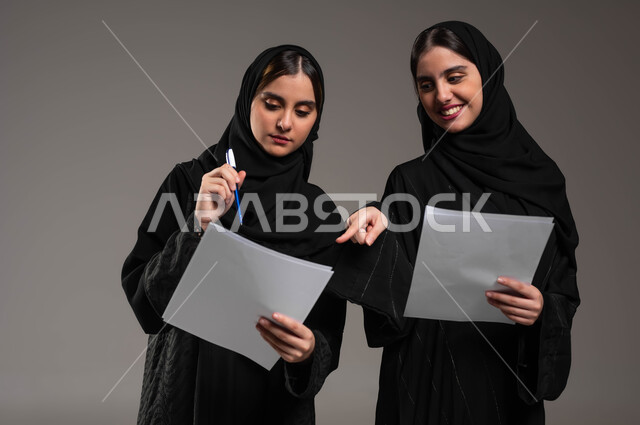 المشاركة والتعاون في إنجاز العمل، ايماءات الفرح والسعادة, مناقشة الاعمال مع فريق العمل, الاشارة بالاصبع على أوراق العمل, بورتريه مقرب لبنتان عربيتان سعوديتان خليجيتان ترتديان الحجاب والعباءة، خلفية رمادية