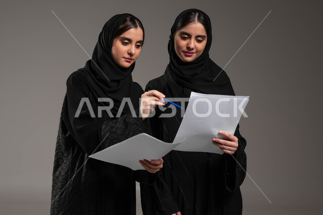 الشراكة والتعاون في انجاز المهام, إعداد الخطط الاستراتيجية، الإشارة إلى أوراق العمل، بورتريه مقرب لبنتان توءم عربيتان سعوديتان خليجيتان ترتديان الحجاب والعباءة تقومان بمناقشة المشاريع، خلفية رمادية